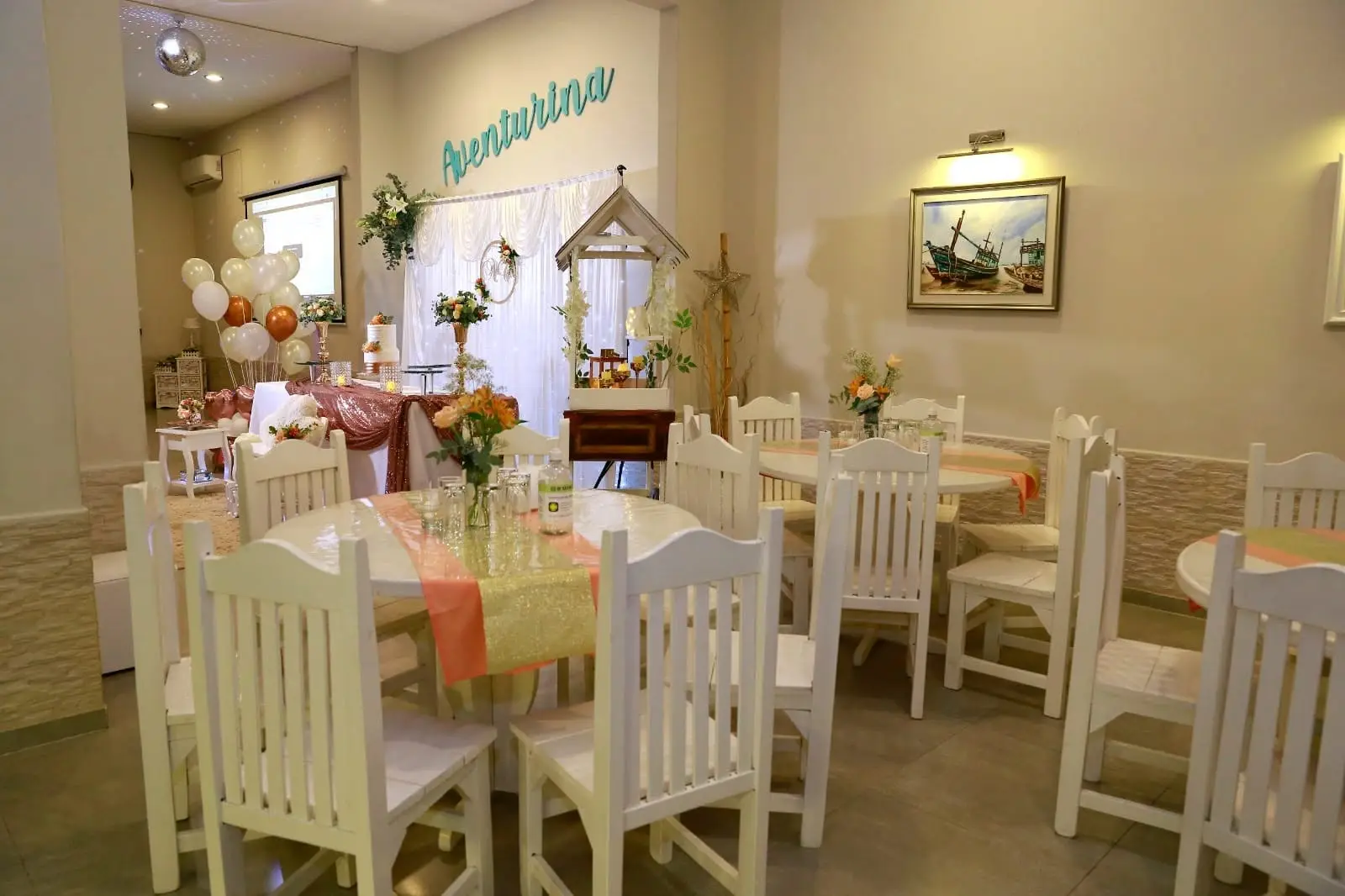 Salón con mesas redondas y sillas blancas de madera, globos y mesa principal al fondo